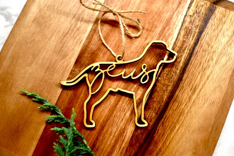 May include: A wooden dog ornament with the name "Zeus" cut out in the design. The ornament is hanging from a piece of twine.