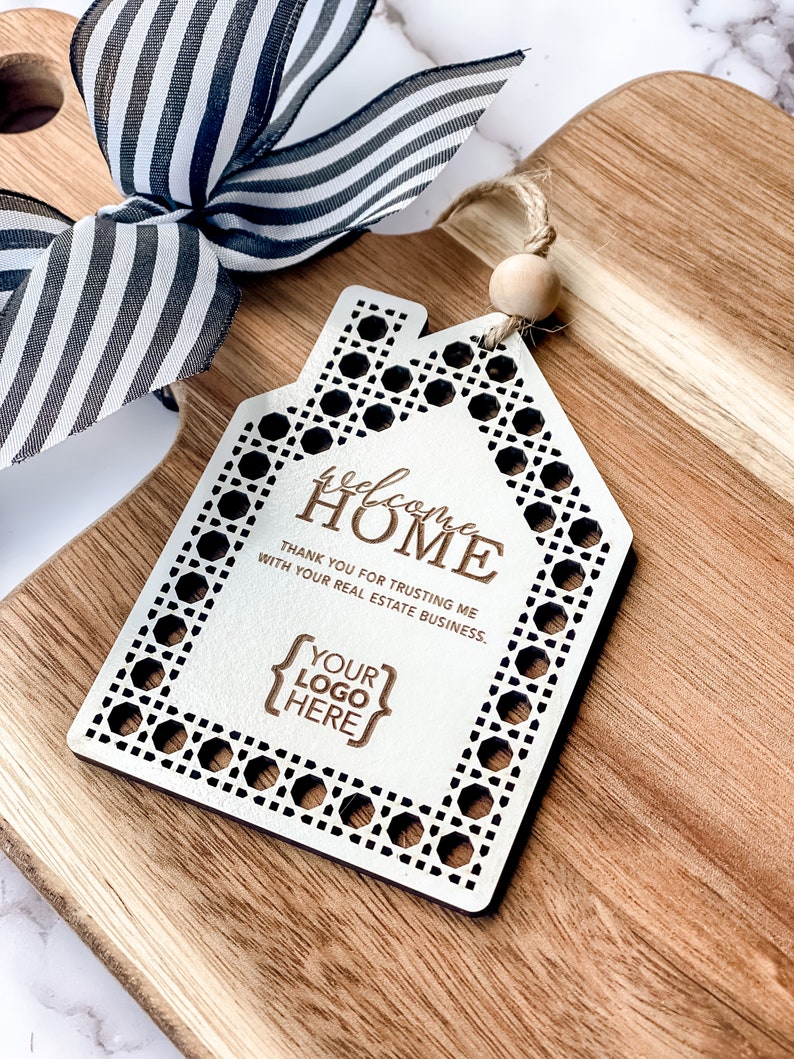 May include: A wooden house-shaped tag with the words "welcome HOME" and "THANK YOU FOR TRUSTING ME WITH YOUR REAL ESTATE BUSINESS." The tag has a decorative border and a space for a logo. A black and white striped ribbon and a wooden bead are also visible.