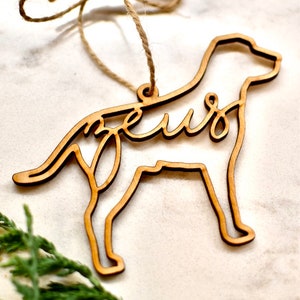 May include: Wooden dog-shaped Christmas ornament with the word "Zeus" cut out in the design. The ornament is brown and has a twine string for hanging.