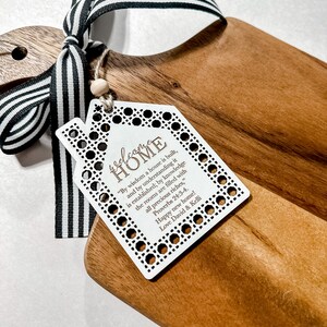 May include: Wooden cutting board with a decorative tag that reads "welcome HOME" and a black and white striped ribbon. The tag has a house shape and includes a Bible verse. A wooden bead is attached to the tag.