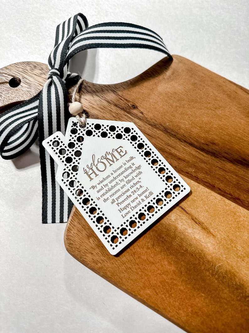 May include: Wooden cutting board with a decorative tag that reads "welcome HOME" and a black and white striped ribbon. The tag has a house shape and includes a Bible verse. A wooden bead is attached to the tag.