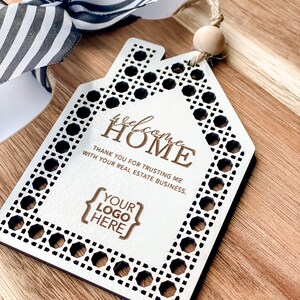 May include: A wooden house-shaped tag with the words "welcome HOME" and "THANK YOU FOR TRUSTING ME WITH YOUR REAL ESTATE BUSINESS." The tag has a decorative border and a space for a logo. It is attached to a wooden cutting board with a black and white striped bow.
