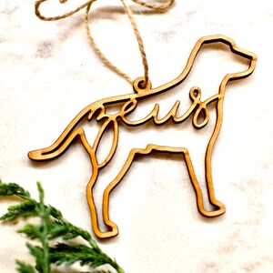 May include: A wooden dog-shaped Christmas ornament with the word "Zeus" cut out in the design. The ornament is hanging from a twine string.