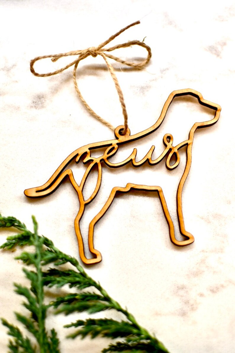 May include: A wooden dog-shaped Christmas ornament with the word "Zeus" cut out in the design. The ornament is hanging from a twine string.