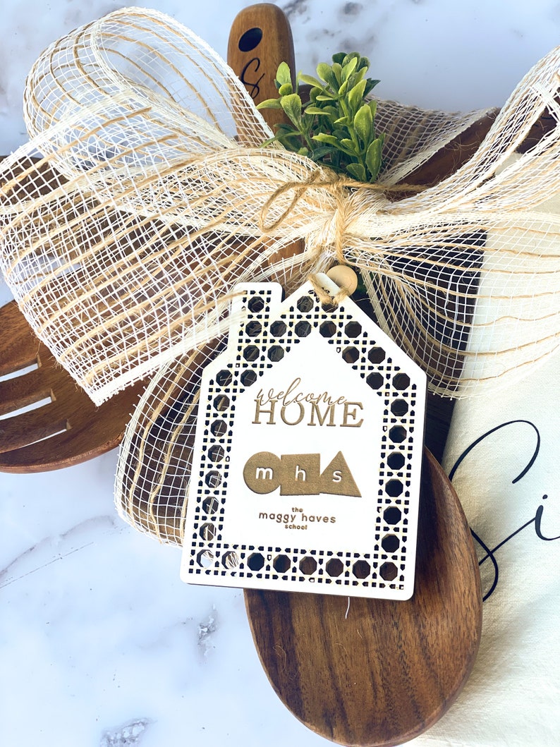 May include: Wooden kitchen utensils, including a spoon and cutting board, are adorned with a decorative bow and a welcome home sign. The sign is a white house shape with the words "welcome home" and "mhs" on it. A sprig of green adds a natural touch.