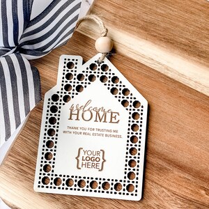 May include: A white wooden house-shaped tag with the words "welcome HOME" and "THANK YOU FOR TRUSTING ME WITH YOUR REAL ESTATE BUSINESS." The tag has a decorative border and a space for a logo. A striped ribbon and wooden cutting board are in the background.