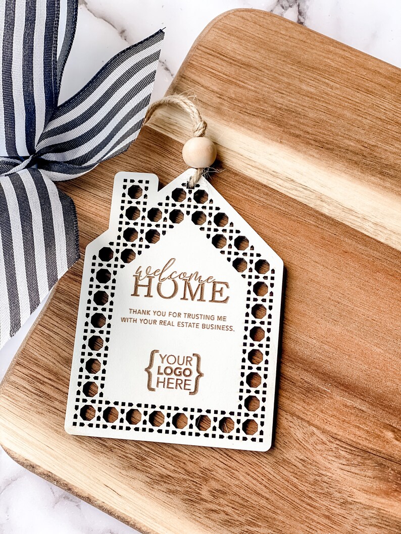May include: A white wooden house-shaped tag with the words "welcome HOME" and "THANK YOU FOR TRUSTING ME WITH YOUR REAL ESTATE BUSINESS." The tag has a decorative border and a space for a logo. A striped ribbon and wooden cutting board are in the background.