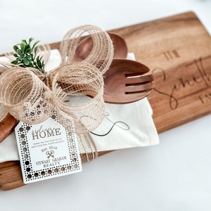 May include: A wooden cutting board with the engraved text "The Smith Family" is paired with wooden utensils, a white cloth, and a decorative bow. A tag reads "welcome HOME" and "STEWART GRAHAM REALTY".