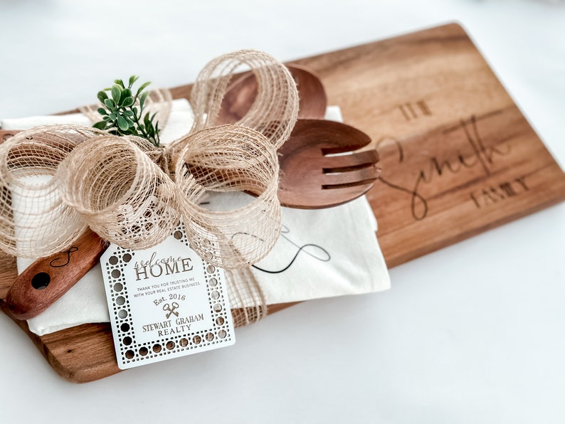 May include: A wooden cutting board with the engraved text "The Smith Family" is paired with wooden utensils, a white cloth, and a decorative bow. A tag reads "welcome HOME" and "STEWART GRAHAM REALTY".