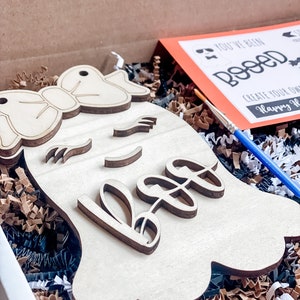 May include: A wooden ghost cutout with a bow and the word "Boo" painted on it. The ghost is in a box with brown and black shredded paper. A paintbrush and a card that says "You've Been Booed! Create Your Own Happy Halloween" are also in the box.