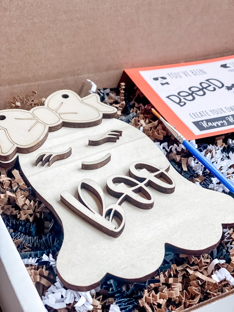 May include: A wooden ghost cutout with a bow and the word "Boo" painted on it. The ghost is in a box with brown and black shredded paper. A paintbrush and a card that says "You've Been Booed! Create Your Own Happy Halloween" are also in the box.