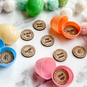 May include: Brightly coloured plastic Easter eggs with wooden tokens inside. The tokens feature phrases such as "No chore day", "You pick dessert", and "Movie Night". Decorative pom-poms are in the background.