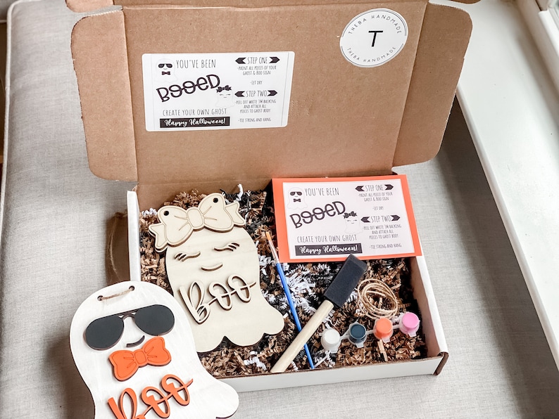 May include: A white box with a brown lid is open, revealing a craft kit for creating your own ghost. The kit includes two wooden ghost shapes, paint, brushes, and instructions. The box is labeled "You've Been Booed!" and "Create Your Own Ghost".