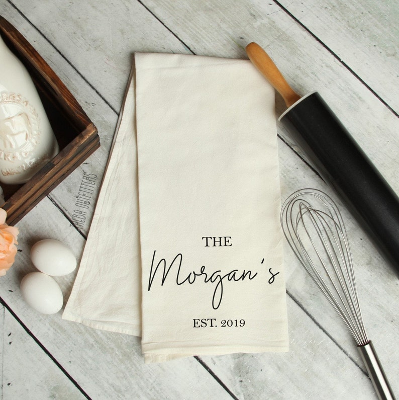 May include: A white kitchen towel with black text that reads "The Morgan's Est. 2019". The towel is lying on a wooden surface with a rolling pin and a whisk.