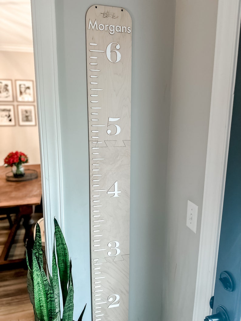 May include: A wooden growth chart with white numbers and the text "The Morgans" at the top. The chart is mounted on a wall and has a light wood finish.