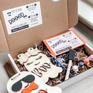 May include: A craft kit for creating two ghost decorations. The kit includes two unfinished wooden ghost shapes, paint, brushes, and instructions. The instructions say "You've Been Booed!" and "Create Your Own Ghost! Happy Halloween!"