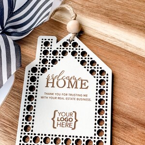 May include: A white wooden house-shaped ornament with a decorative border and the words "Welcome Home" engraved. The ornament includes the text "Thank you for trusting me with your real estate business" and space for a logo. A black and white striped ribbon is visible.