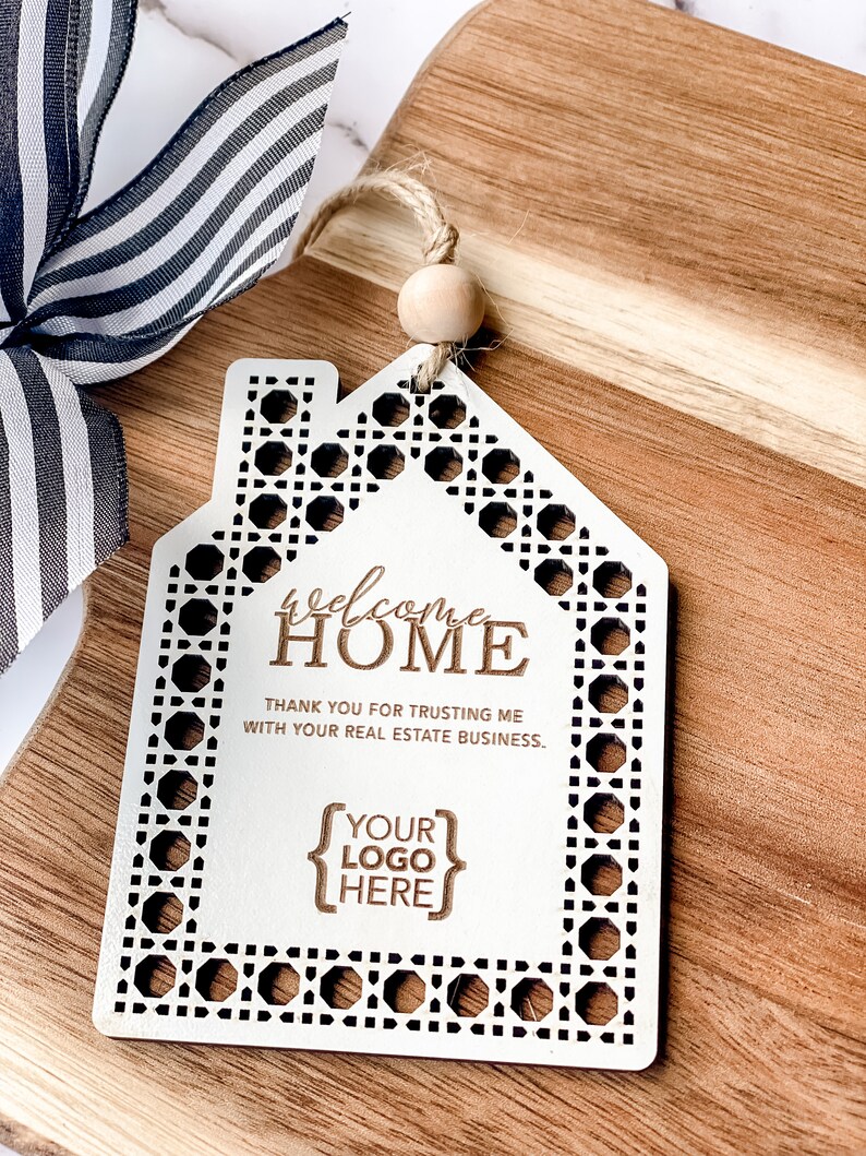 May include: A white wooden house-shaped ornament with a decorative border and the words "Welcome Home" engraved. The ornament includes the text "Thank you for trusting me with your real estate business" and space for a logo. A black and white striped ribbon is visible.