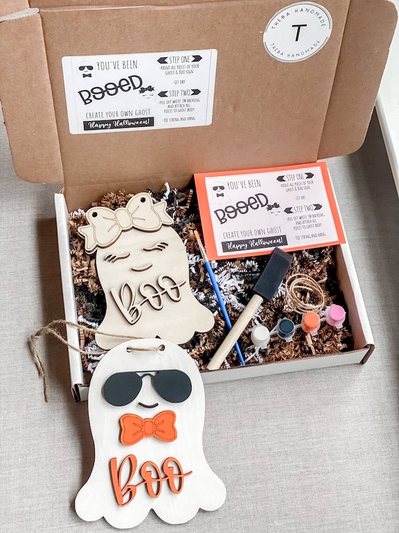 May include: A DIY craft kit to create a ghost decoration. The kit includes a wooden ghost cutout, paint, a paintbrush, twine, and instructions. The ghost has a bow, sunglasses, and the word "Boo" written on it. The instructions say "You've Been Booed!" and "Create Your Own Ghost! Happy Halloween!"