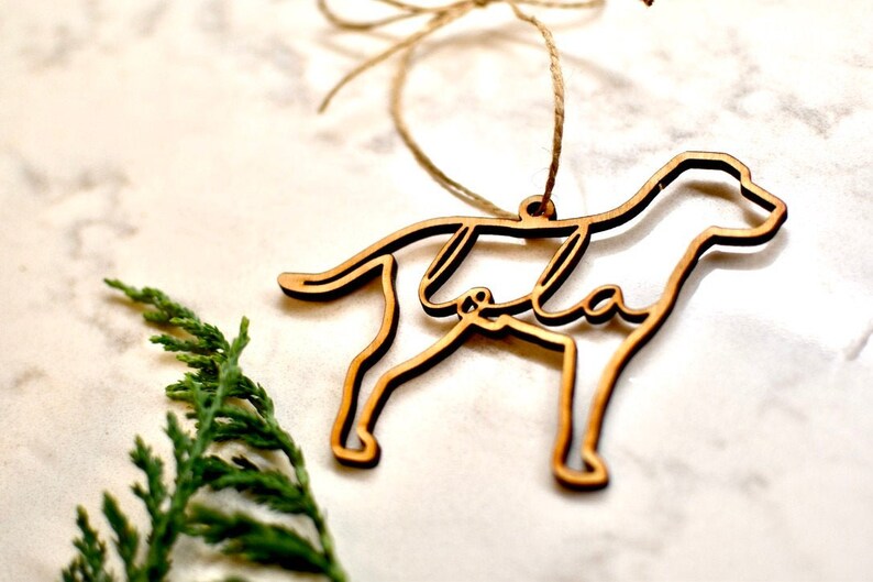 May include: A wooden dog ornament with the name "Lola" cut out inside the silhouette. The ornament is hanging from a twine string.