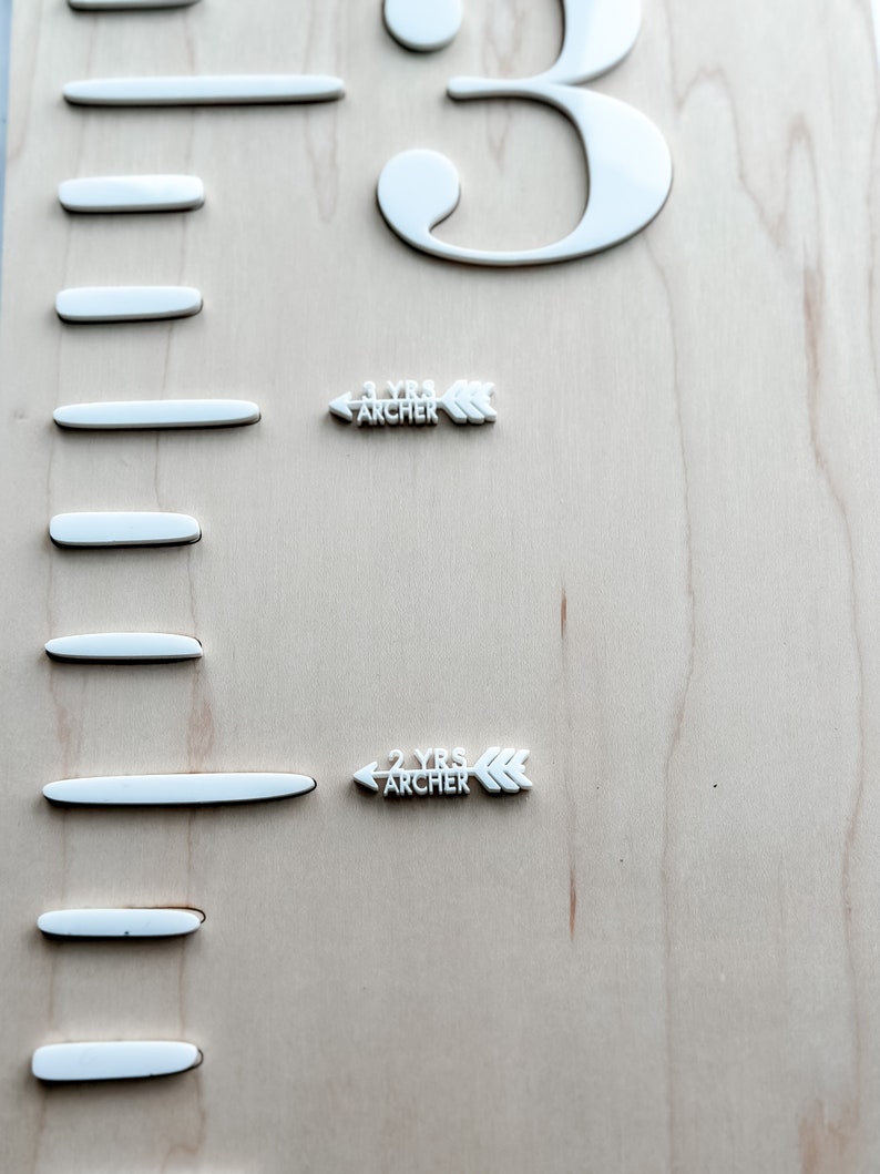 May include: A white wooden growth chart with white markings and a white number "3" at the top. There are two white arrows with the text "1 YRS ARCHER" and "2 YRS ARCHER" on the chart.