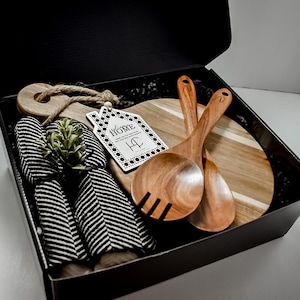 May include: A gift box containing a wooden cutting board, salad servers, striped napkins, and a decorative plant. The cutting board has a round handle and a tag that says "HOME". The box is black.