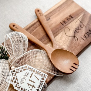 May include: Wooden serving spoons and a cutting board with the words "HOME Sweet HOME" engraved. A decorative tag with the text "welcome HOME 417" is attached with a ribbon. The spoons and board are made of light brown wood.