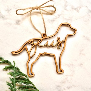 May include: A wooden dog-shaped Christmas ornament with the word "Zeus" cut out inside the silhouette. The ornament is hanging from a twine string.