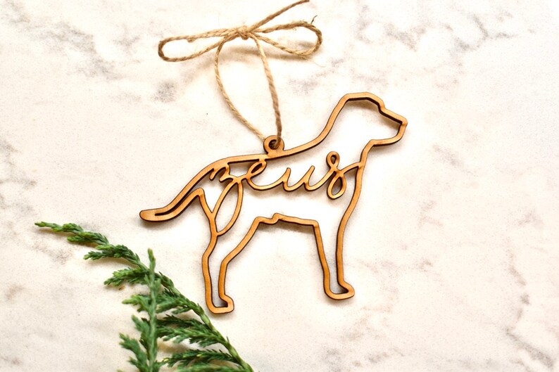 May include: A wooden dog-shaped Christmas ornament with the word "Zeus" cut out inside the silhouette. The ornament is hanging from a twine string.