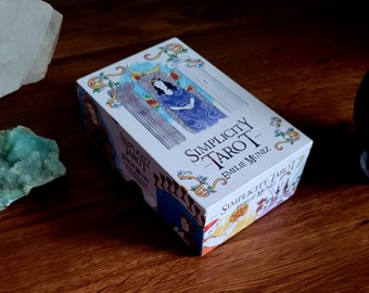 May include: A white box with a colorful illustration of a tarot card on the front. The box is titled "Simplicity Tarot" with the name "Emilie Muniz" printed below.
