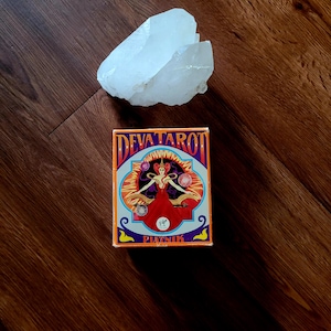 May include: A clear quartz crystal sits next to a tarot card box. The box is decorated in orange, blue, and purple, with the text "DEVA TAROT" and "PIATNIK". The box features an illustration of a woman in a red dress.
