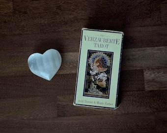 Puede incluir: Una piedra de selenita blanca en forma de corazón junto a una caja rectangular de cartas de tarot titulada Das Verzauberte Tarot de Amy Zerner y Monte Farber, con una colorida ilustración de una luna creciente y un paisaje en la portada.