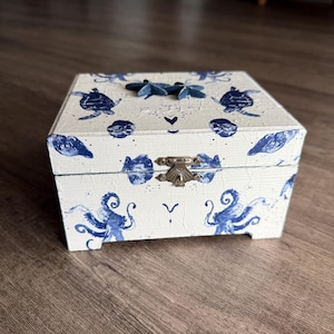 May include: A white wooden box with blue sea creature designs, including turtles, starfish, and octopuses. The box has a metal clasp and is decorated with blue accents. The box is sitting on a wooden surface.
