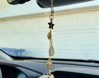 May include: A gold-toned metal chain hanging from a vehicle rearview mirror, featuring a series of charms including a dark starfish, small metallic beads, and several natural seashells, ending with a large, fan-shaped white scallop shell.