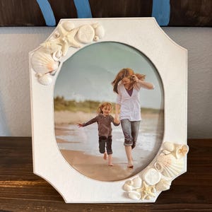 May include: White, octagonal picture frame with a beach theme. The frame is decorated with seashells and starfish. The frame holds a photo of a woman and child running on a beach.