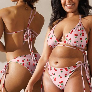 May include: Pink bikini set with a cherry pattern. The two-piece swimsuit features a triangle top and a low-rise bottom, both adorned with red cherries and green leaves. The bikini has tie closures for an adjustable fit.