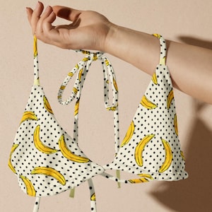 Può includere: Top bikini bianco con motivo a banane gialle e pois neri. Il top a triangolo ha lacci da annodare intorno al collo e sulla schiena. Il top del bikini è tenuto da una mano.