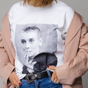 May include: White t-shirt with a black and white portrait graphic. The person is wearing a light pink blazer, blue jeans, and a black belt with a silver buckle. The person has short, light brown hair.