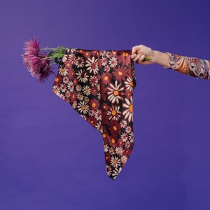 May include: A burgundy floral patterned scarf with white and orange accents is held up by a hand, with purple flowers on the left. The scarf is against a purple background. The sleeve of the person holding the scarf is patterned.