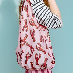 Puede incluir: Una mujer con una camisa a rayas rosa y blancas con estampado de langosta, pantalones a rayas rosa y blancas, botas marrones y un sombrero de mezclilla azul con borde deshilachado. Lleva gafas de sol y tiene la mano en la barbilla.