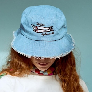 May include: A woman wearing a white t-shirt, pink striped trousers, and a blue denim bucket hat with an embroidered design of two fish and the text "La Sardine by Marvilla".