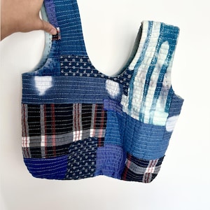 Könnte beinhalten: Eine Patchwork-Tragetasche in verschiedenen Blautönen mit weißen und roten Akzenten. Die Tasche hat ein gestepptes Design mit verschiedenen Stoffmustern, darunter Streifen, Tupfen und Karos. Die Tasche wird von einer Hand gehalten.