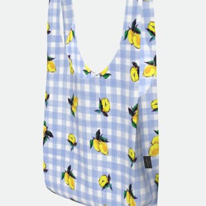 May include: A reusable shopping bag with a blue and white gingham pattern, adorned with yellow lemons and green leaves. The bag has two handles and a small black tag.