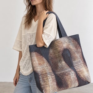 Op de afbeelding: Een grote, rechthoekige draagtas van donkerblauwe denim met een opvallend, abstract golvend patroon in bruin-, beige- en wittinten. De tas heeft twee schouderbanden en is voorzien van zichtbare, dichte horizontale stiksels over het gehele oppervlak.