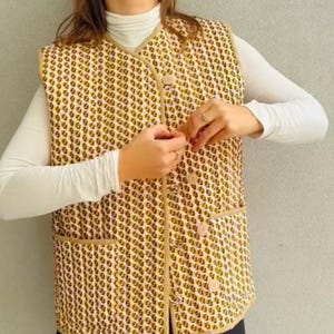 Może przedstawiać: Bezrękawnik z żółto-różowym geometrycznym wzorem. Kamizelka zapinana na guziki, z kieszenią z przodu. Osoba nosząca kamizelkę ma na sobie również białą koszulę z golfem, czarną spódnicę i czarne rajstopy.