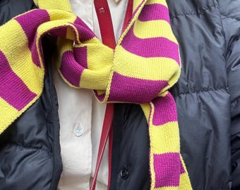 May include: A knitted scarf featuring horizontal stripes in bright yellow and magenta. The scarf is worn over a cream-colored button-down shirt and a black quilted jacket, with a thin red strap visible hanging vertically in the center.