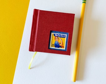 May include: A small red notebook with a vintage "We Can Do It!" stamp featuring a woman with a red bandana and blue shirt. The notebook has a yellow ribbon attached to it and is sitting on a white surface next to a yellow pencil.