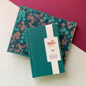 May include: A teal green journal with the text "No Robots Here" on a white sticker. The journal has a floral pattern on the cover and is made with heart and hands, not gaskets and gears.