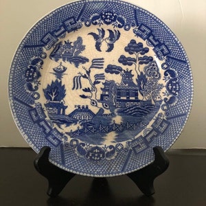 May include: A blue and white Willow pattern plate. The plate features a detailed landscape scene with pagodas, bridges, and birds. The rim has a decorative border. The plate is displayed on a black stand.