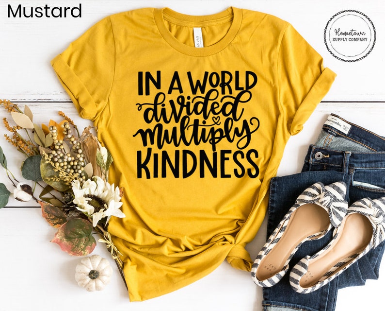 May include: A mustard yellow t-shirt with the text "In a world divided multiply kindness" in black lettering. The shirt is laid flat on a white wooden surface with a bouquet of flowers and a pair of jeans and shoes.